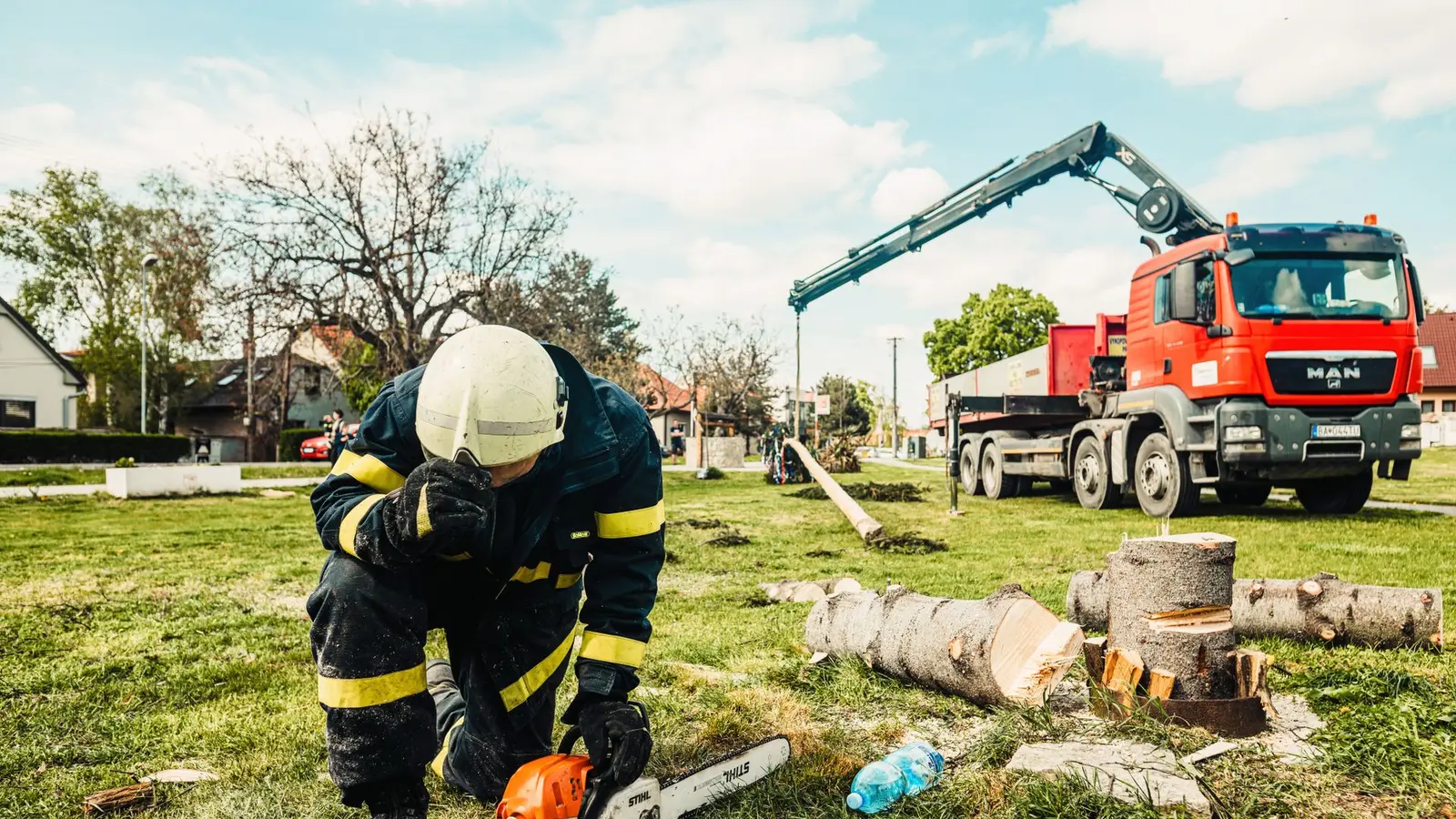 Worker in a helmet cutting logs with a chainsaw near a truck-mounted crane
